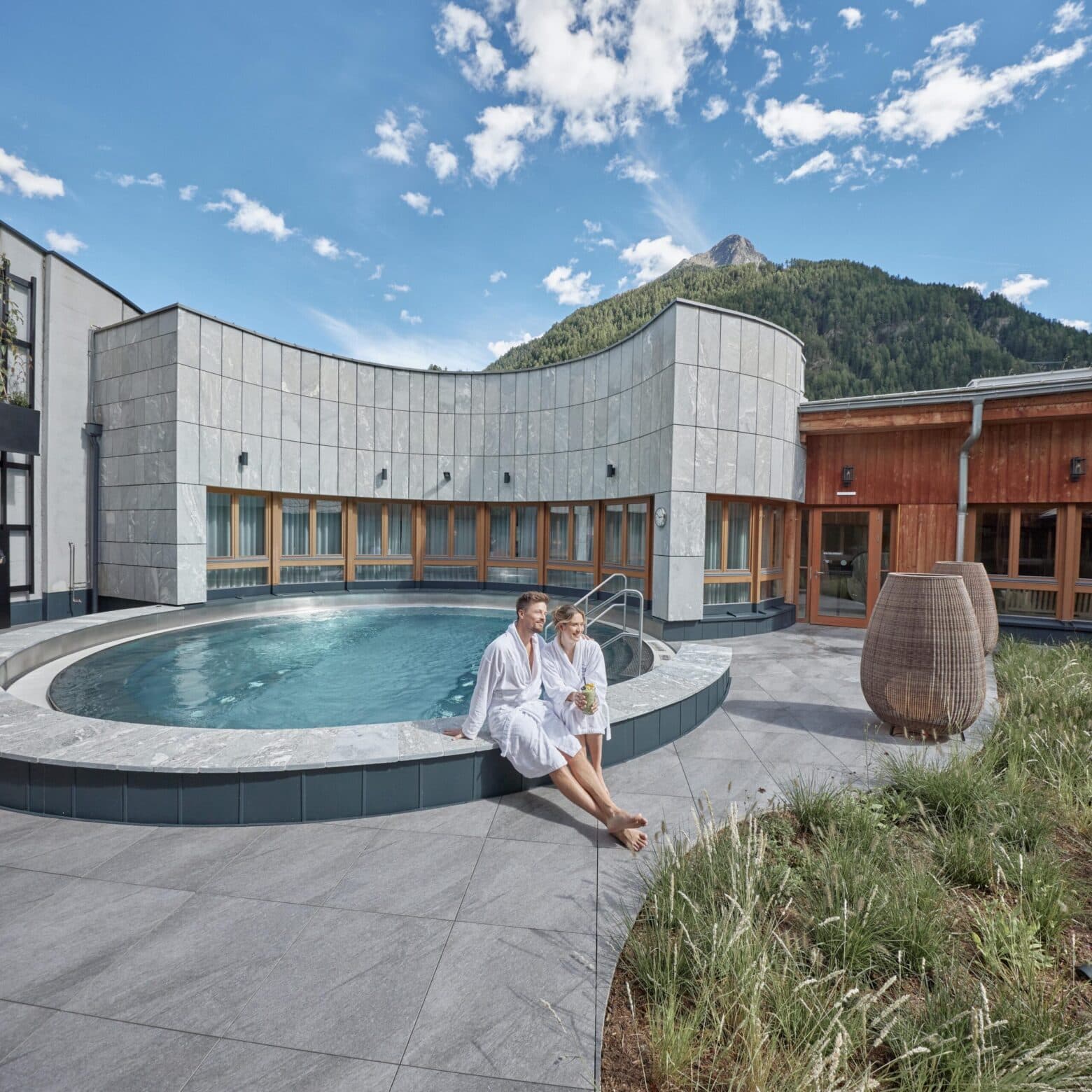 Zwei Personen in weißen Gewändern sitzen an einem kreisrunden Außenpool mit einem Berg und blauem Himmel im Hintergrund, umgeben von modernen Gebäuden und Grünanlagen.