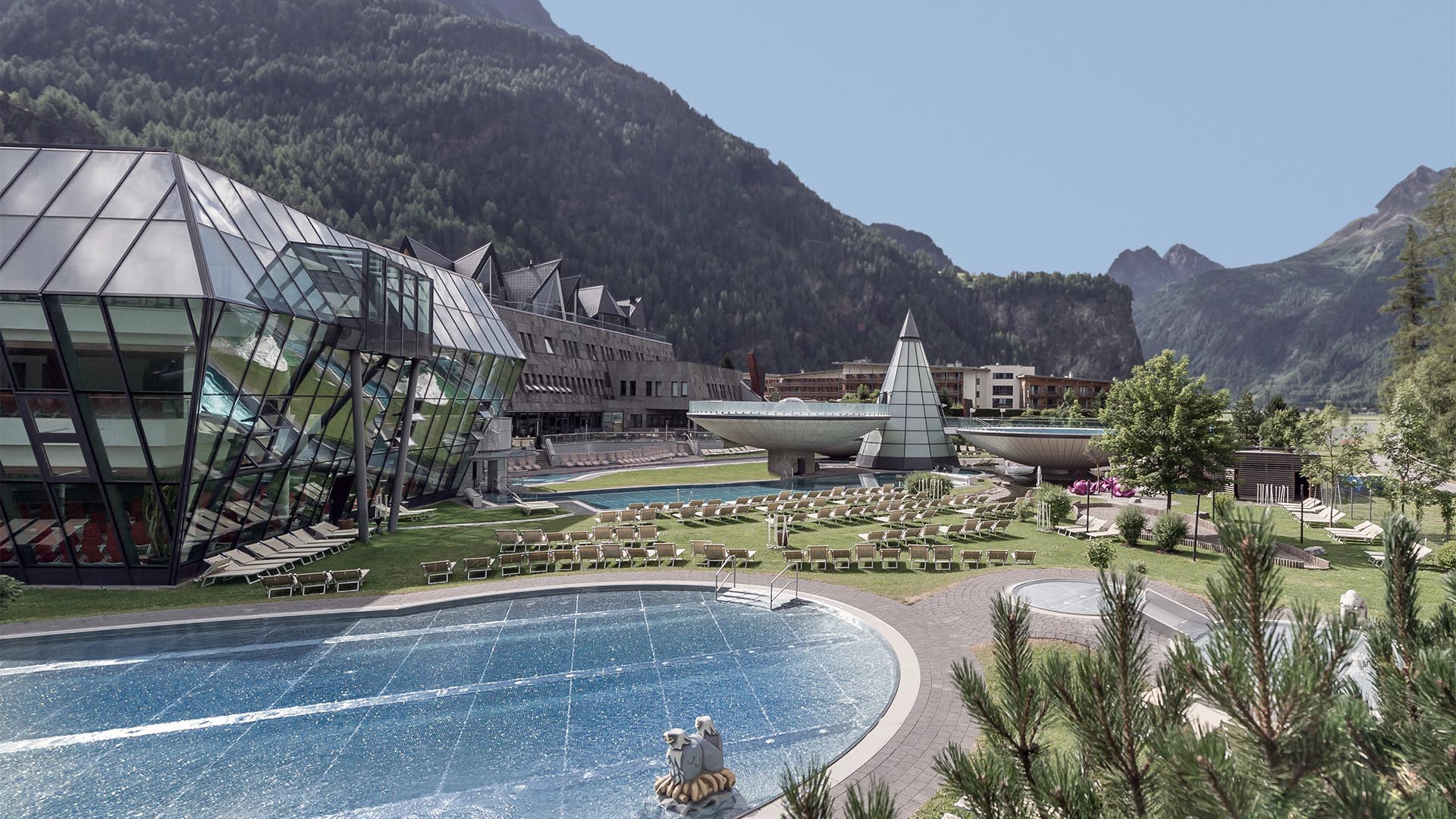 Modernes Spa-Resort mit Glasarchitektur, Außenpools und Liegestühlen vor der Kulisse grüner Berge und klarer Himmel.