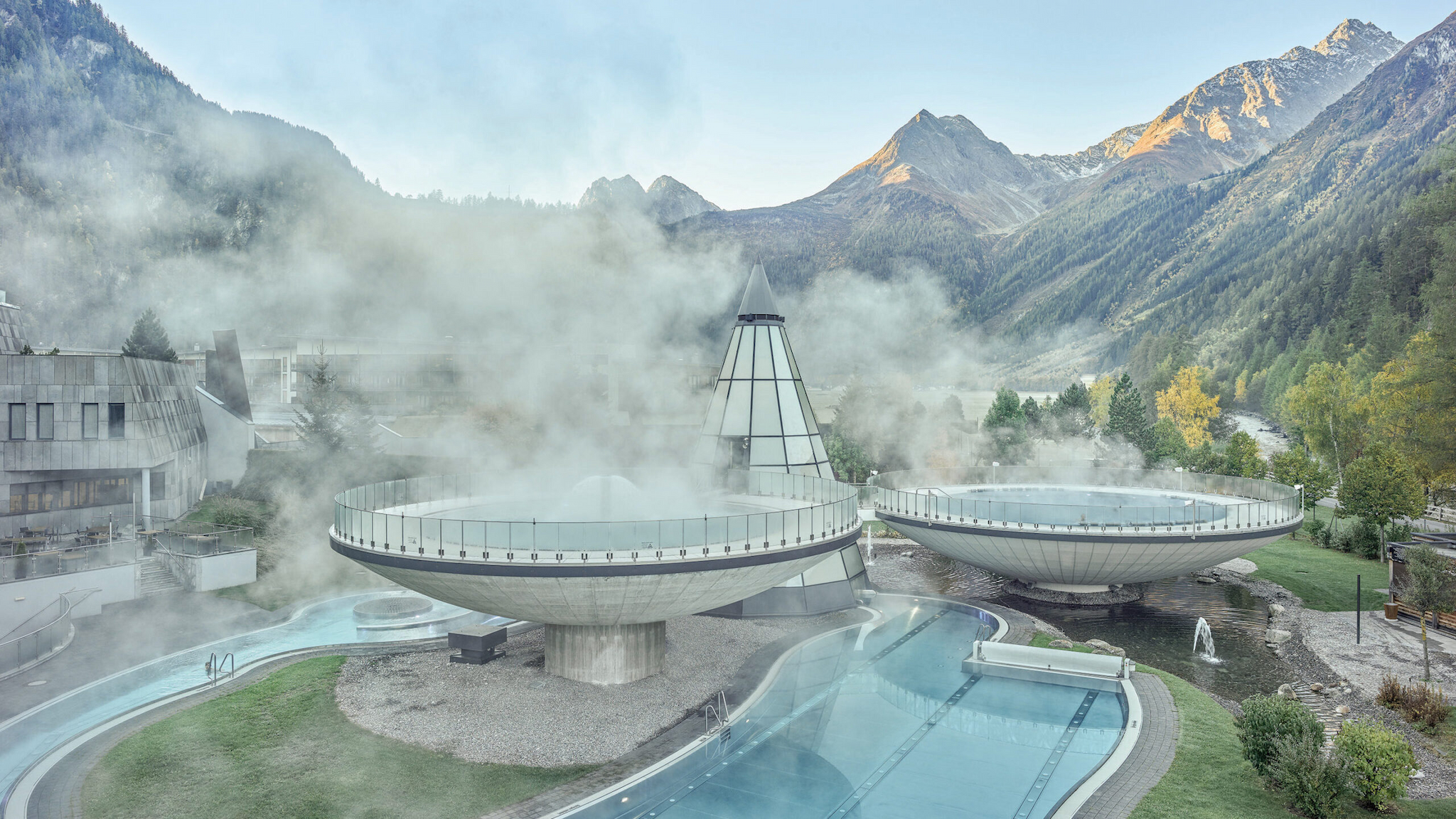 Futuristic buildings with steam rising leave an indelible imprint on the landscape, featuring large round structures and a central conical tower set amidst mountainous greenery.