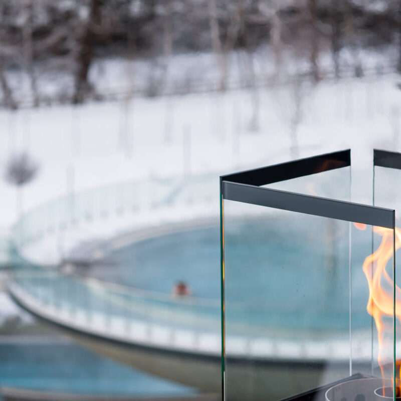 Im Vordergrund brennt eine verglaste Flamme, im Hintergrund ist eine verschwommene Ansicht einer Person zu sehen, die in einem runden Außenpool schwimmt, der von Schnee umgeben ist.