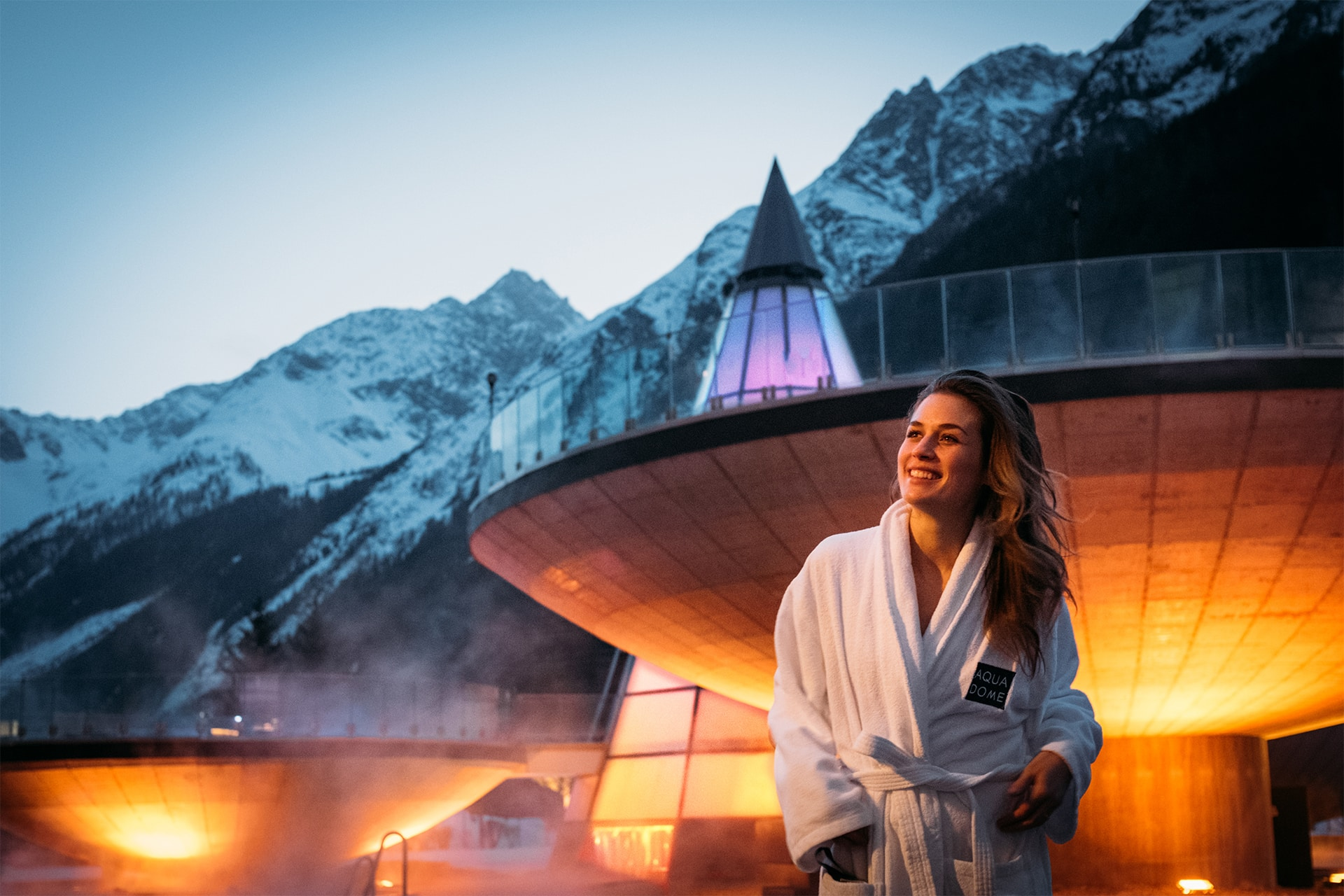 Person in a white robe stands in front of a modern building with mountains in the background at dusk.