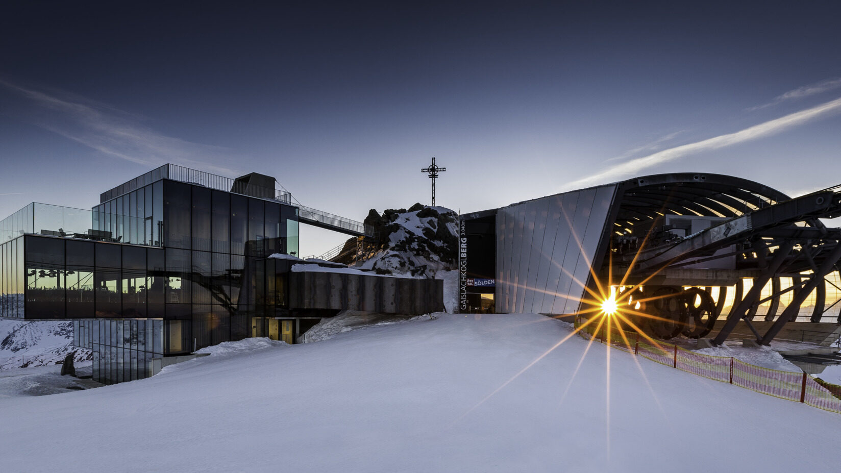 Une station de montagne moderne en verre et métal avec un environnement enneigé, présentant un soleil couchant partiellement visible à travers une ouverture.