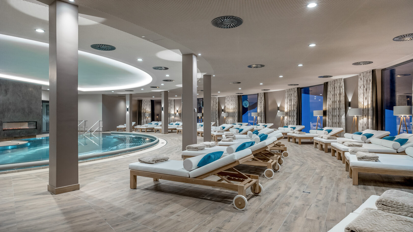 Luxuriöses Spa-Interieur mit rundem Innenpool, umgeben von Holzliegen mit weißen Kissen und gerollten Handtüchern. Sanfte Beleuchtung und modernes Design schaffen eine entspannende Atmosphäre.
