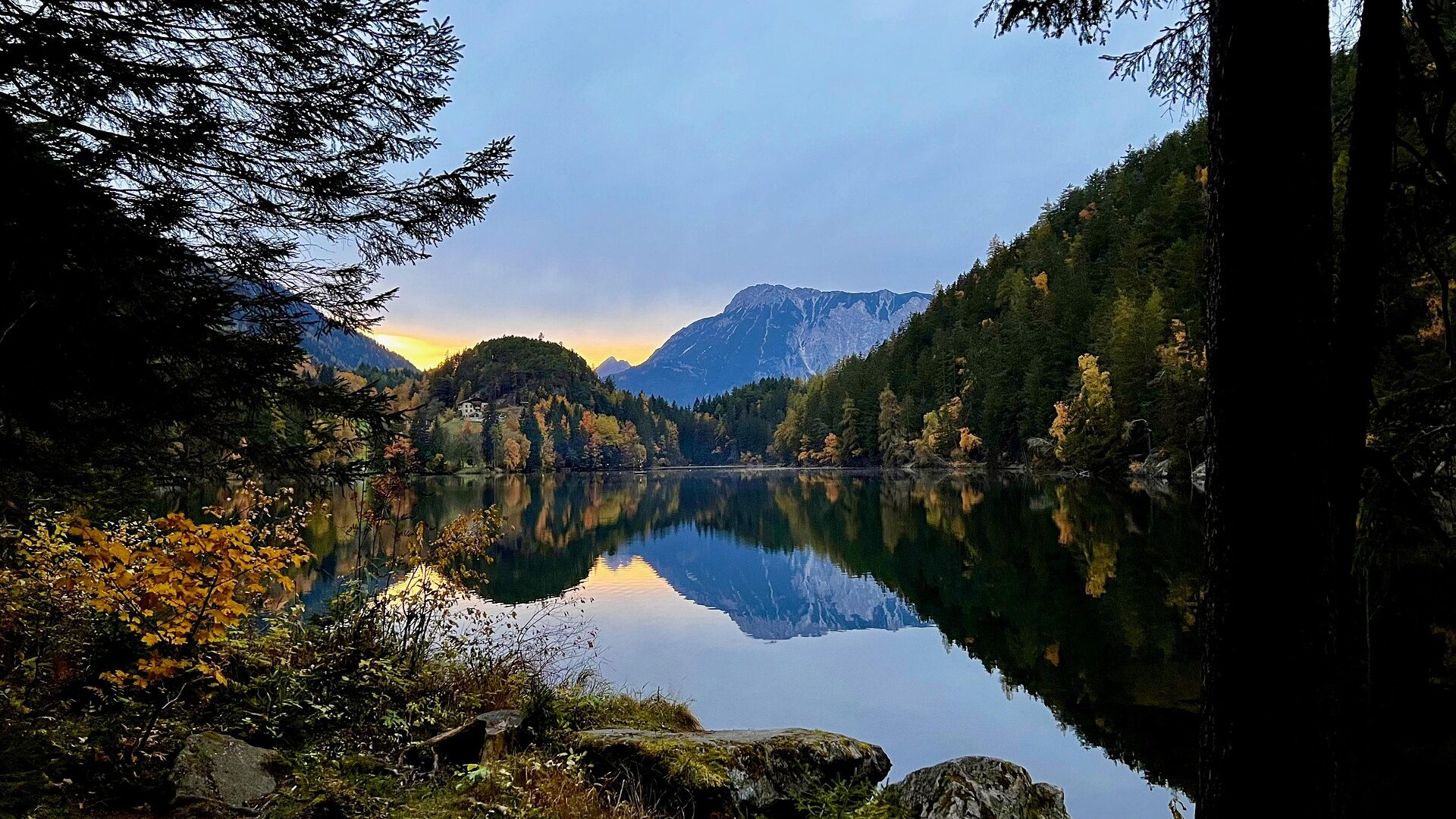 Ein ruhiger See, umgeben von Bäumen und Bergen, spiegelt die Landschaft bei Sonnenuntergang, mit Herbstlaub und einem teilweise bewölkten Himmel.