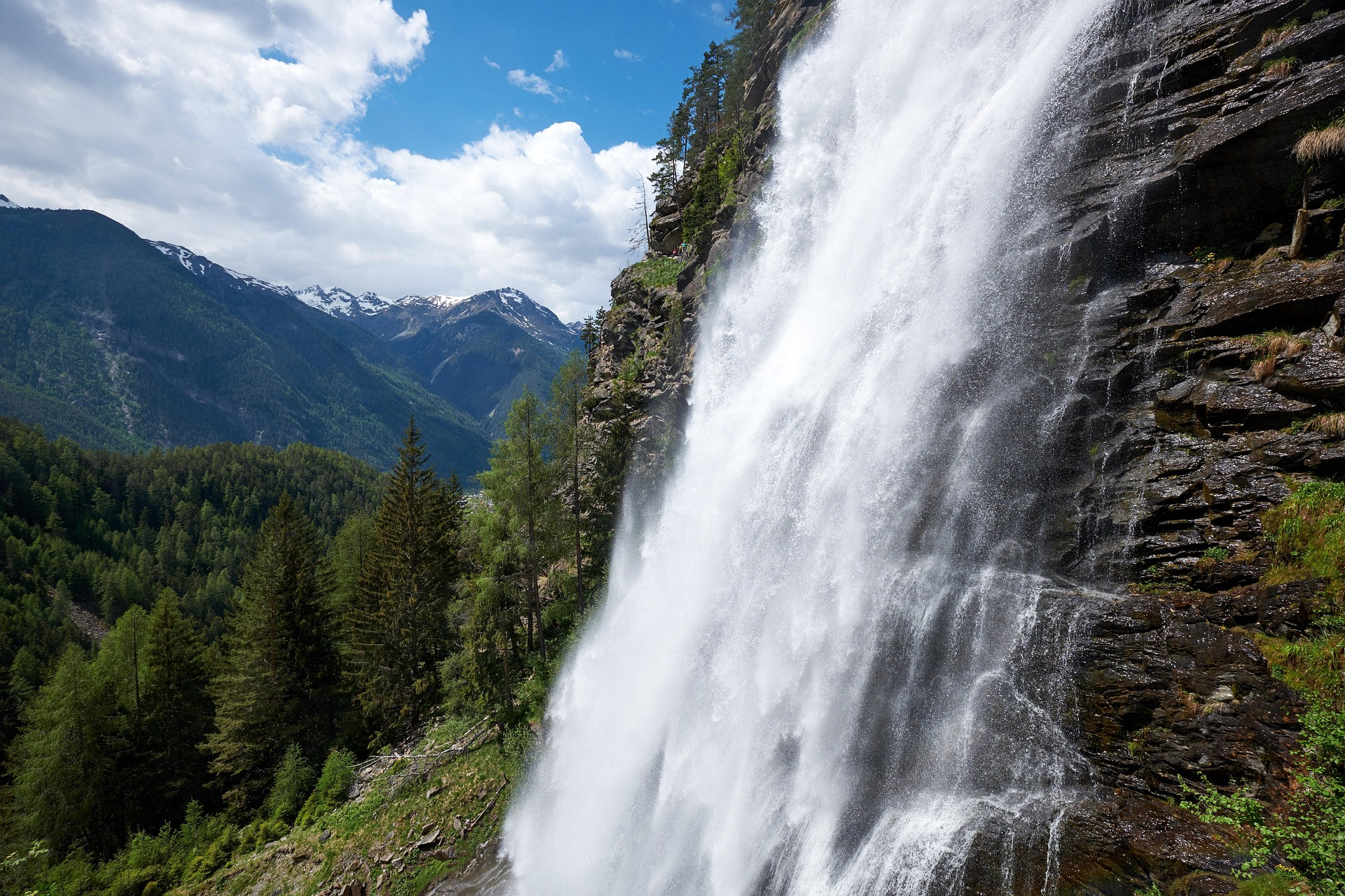 Ein Wasserfall stürzt über eine felsige Klippe, umgeben von grünem Wald, mit schneebedeckten Bergen und einem teilweise bewölkten Himmel im Hintergrund.