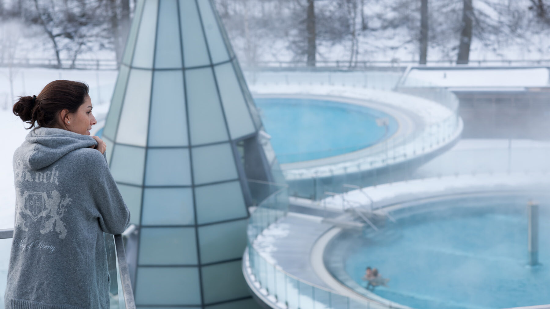 A woman in a gray sweater stands overlooking steaming outdoor pools surrounded by snow, with people swimming in the water.