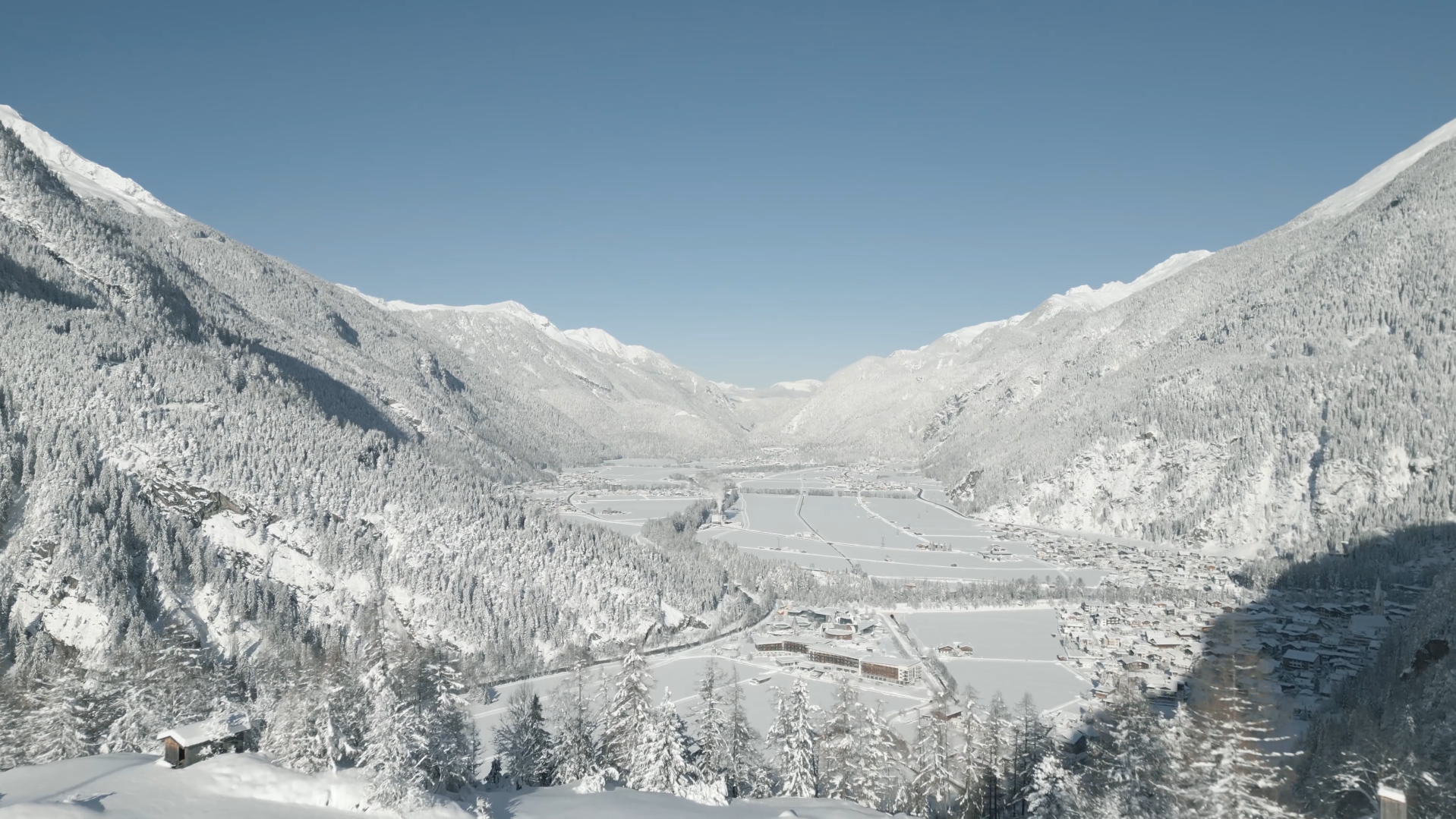 A snow-covered valley surrounded by tall mountains under a clear blue sky, with a small village and forested slopes in the foreground.