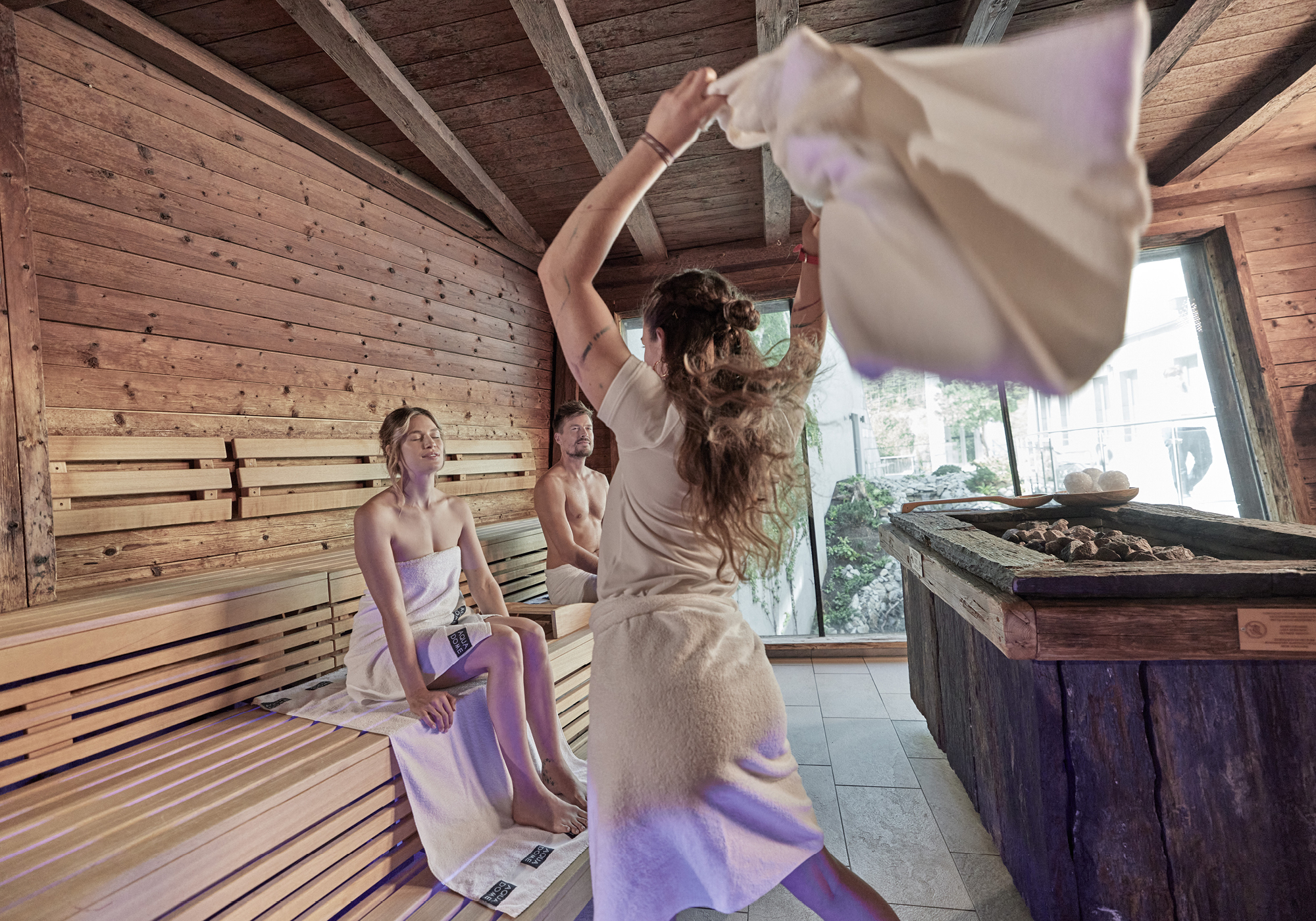 Three people in towels are in a wooden sauna; one person is standing and waving a towel, while two others sit on a bench and watch.