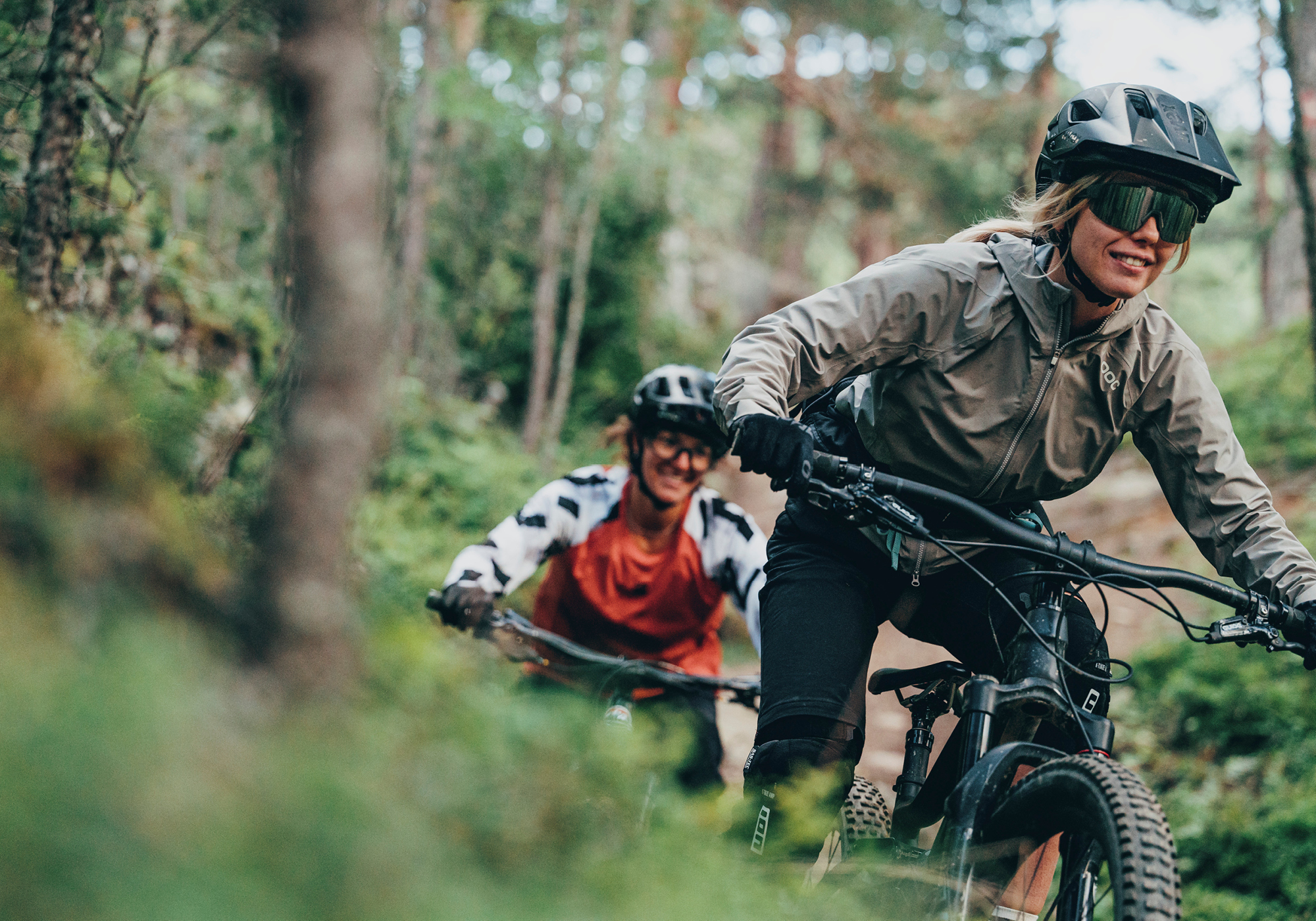 Due persone in mountain bike su un sentiero forestale, entrambe con casco e occhiali da sole. La persona davanti sorride, seguita da vicino da un altro ciclista.