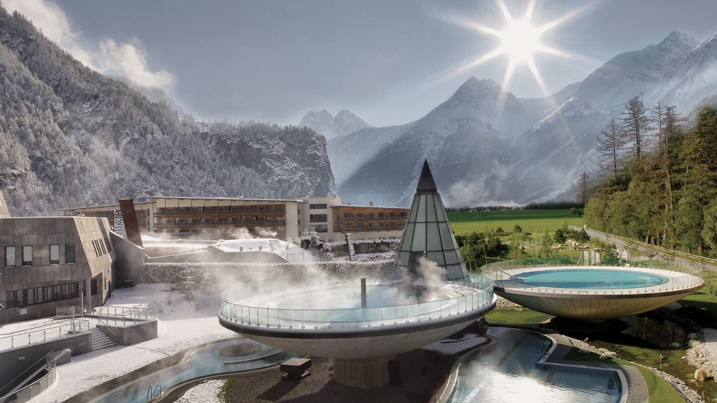 Ein modernes alpines Resort mit kreisrunden Außenbecken, eines davon mit aufsteigendem Dampf, vor der Kulisse verschneiter Berge und einer strahlenden Sonne am klaren Himmel.
