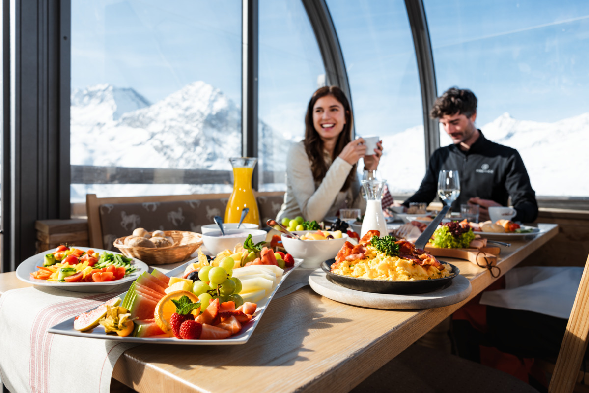 A couple enjoys breakfast at a table with fruit, scrambled eggs, pastries, and juice in a sunlit restaurant with snowy mountains visible through large windows.