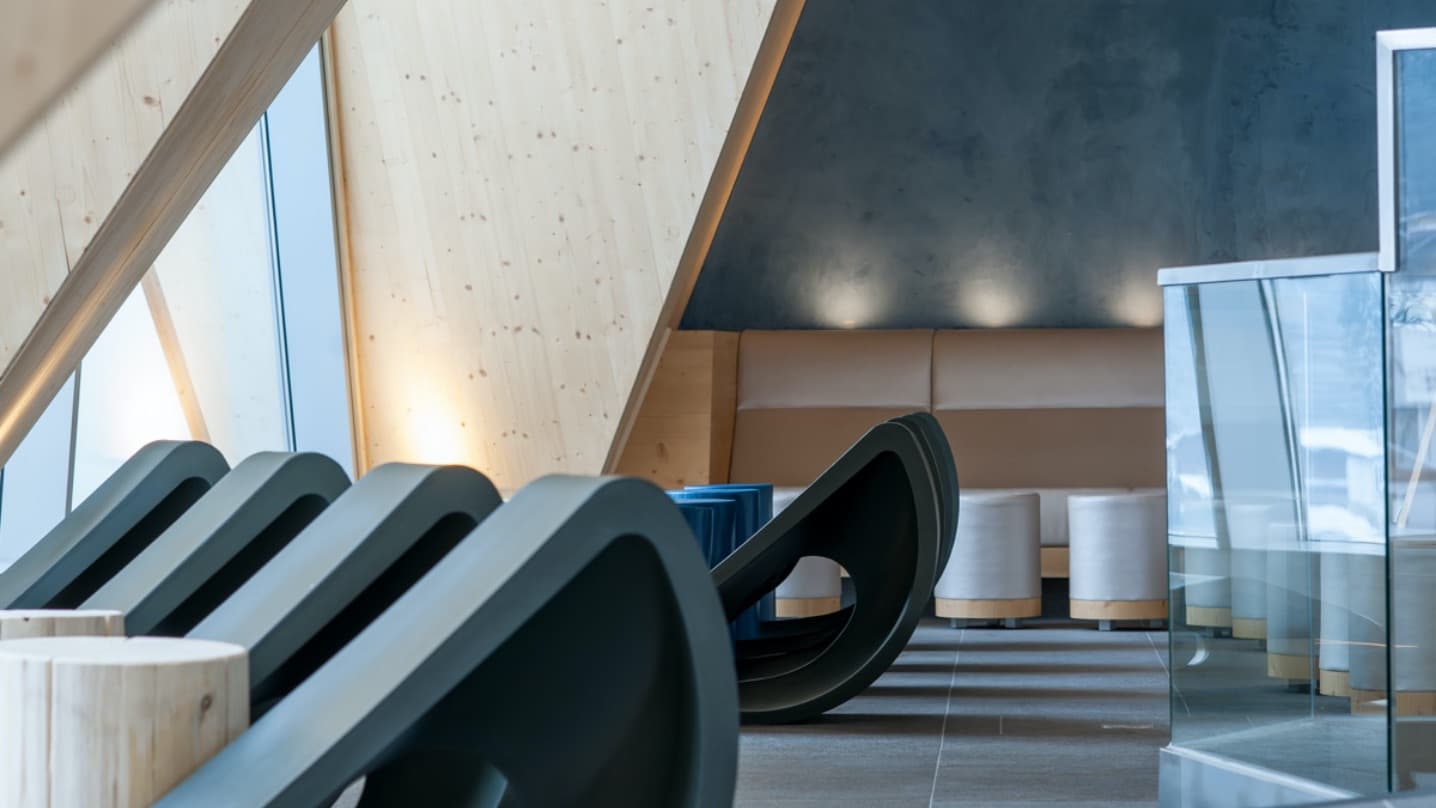 Modernes Interieur mit abgewinkelten Holzbalken, eleganten schwarzen Stühlen, einer beige gepolsterten Bank, zylindrischen Hockern und einer Glastrennwand auf der rechten Seite, die einen zeitgenössischen Sitzbereich schafft.