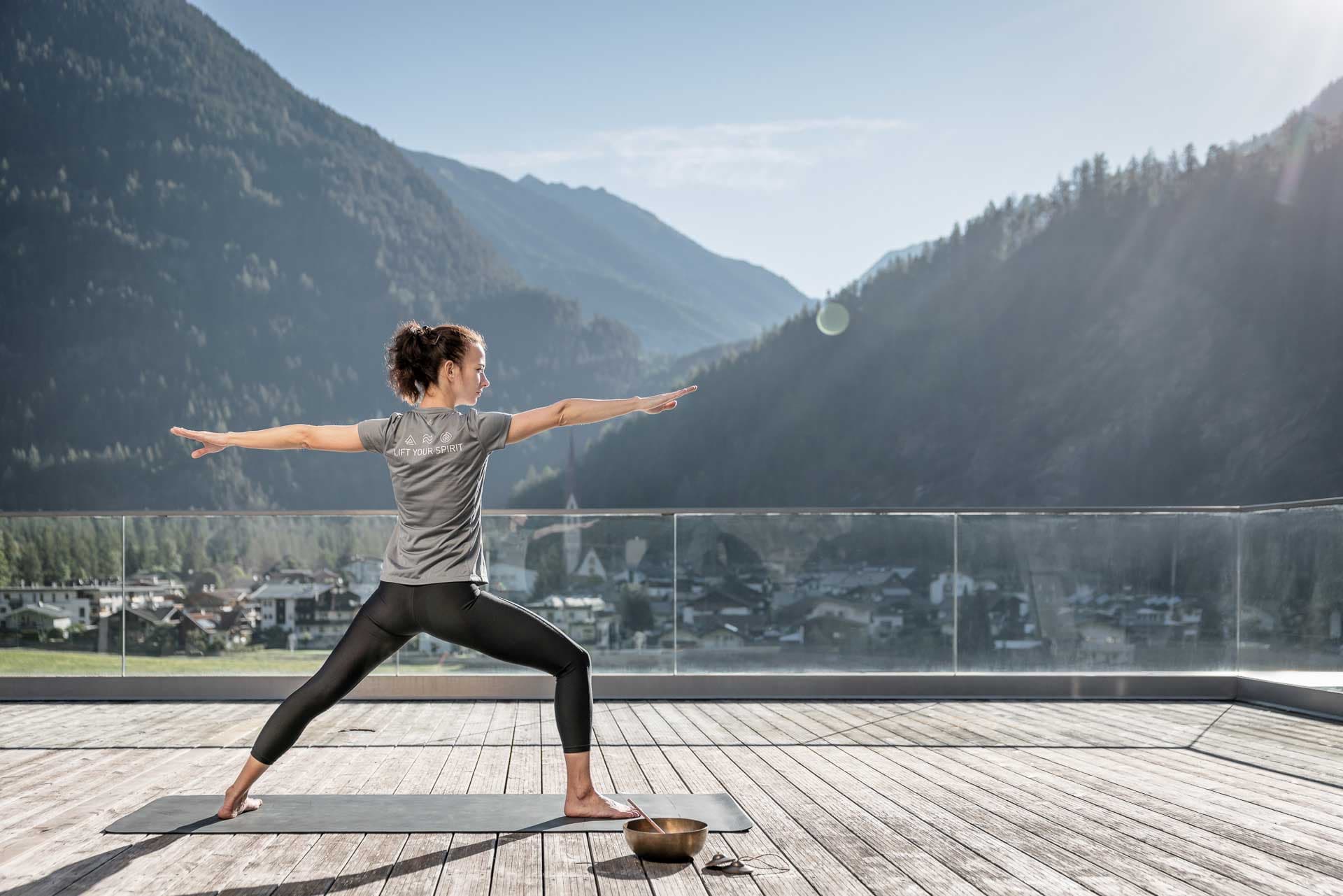 Person, die Yoga in der Krieger-II-Pose auf einem Holzdeck mit Bergen im Hintergrund praktiziert.