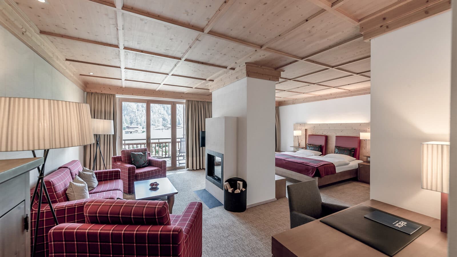 This cozy hotel room features a warm seating area with red plaid couches, a charming fireplace, and a comfortable bed. A wooden ceiling and large windows reveal an inviting view outside, while neutral tones and soft lighting enhance the tranquil atmosphere.
