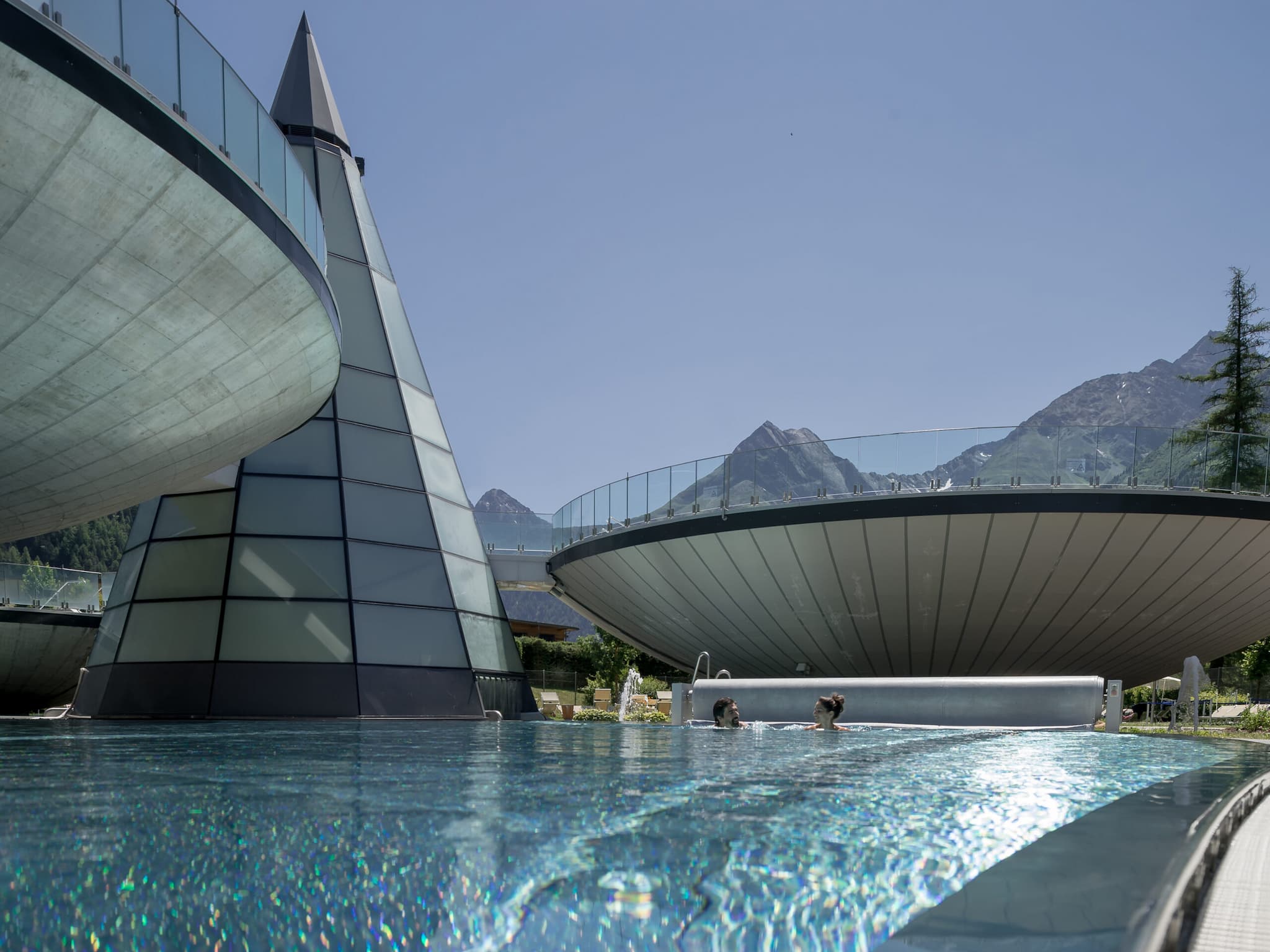Futuristic pools with a stunning glass pyramid and circular structures, where two people swim, all set against a backdrop of majestic mountains.