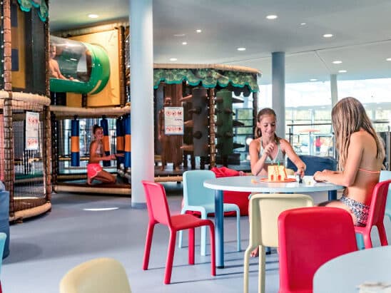 Des enfants jouent dans une aire de jeux intérieure ; deux filles sont assises à une table avec des jouets, tandis qu'un autre enfant est dans une structure de jeu avec des toboggans et des sections d'escalade.