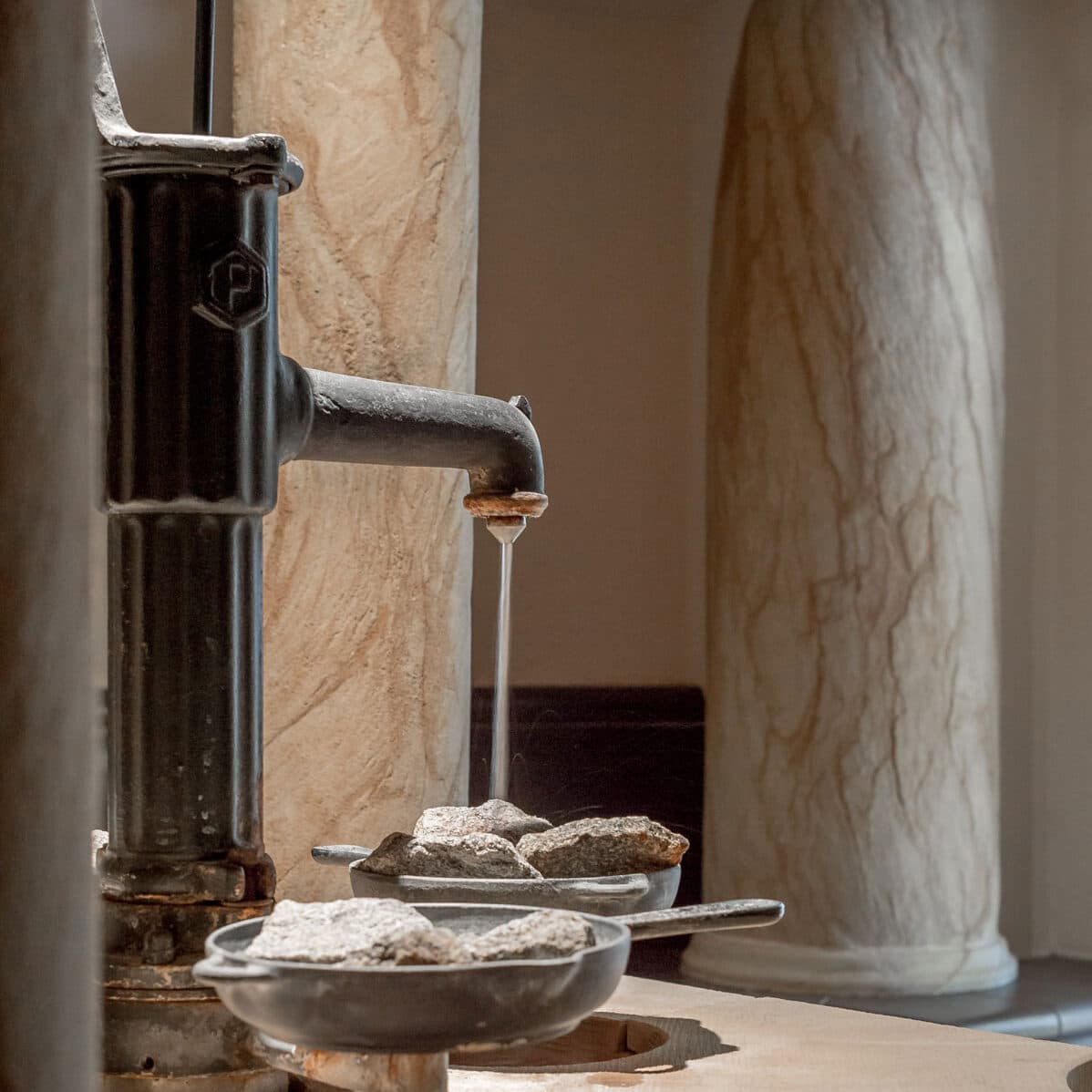 Wasser fließt aus einem Metallausguss in mit Steinen gefüllte Schalen, die von Marmorsäulen umgeben sind und so die ruhige Atmosphäre einer luxuriösen Sauna hervorrufen.