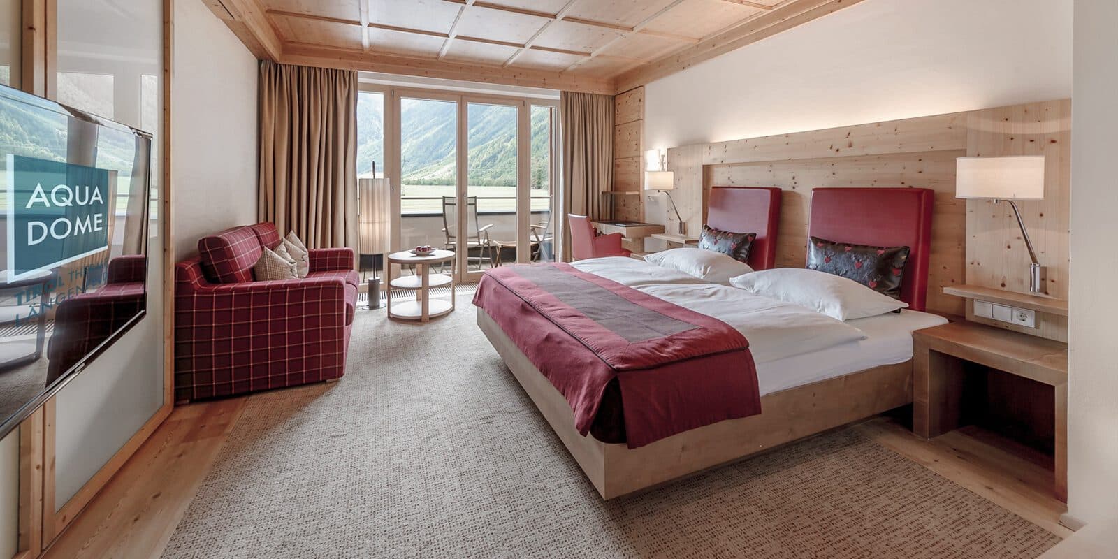 Hotelzimmer mit Doppelbett, Holzdekor und Sofa. Großes Fenster mit Blick auf die Berge. Fernseher an der gegenüberliegenden Wand und kleiner Balkon mit Sitzgelegenheiten im Freien.