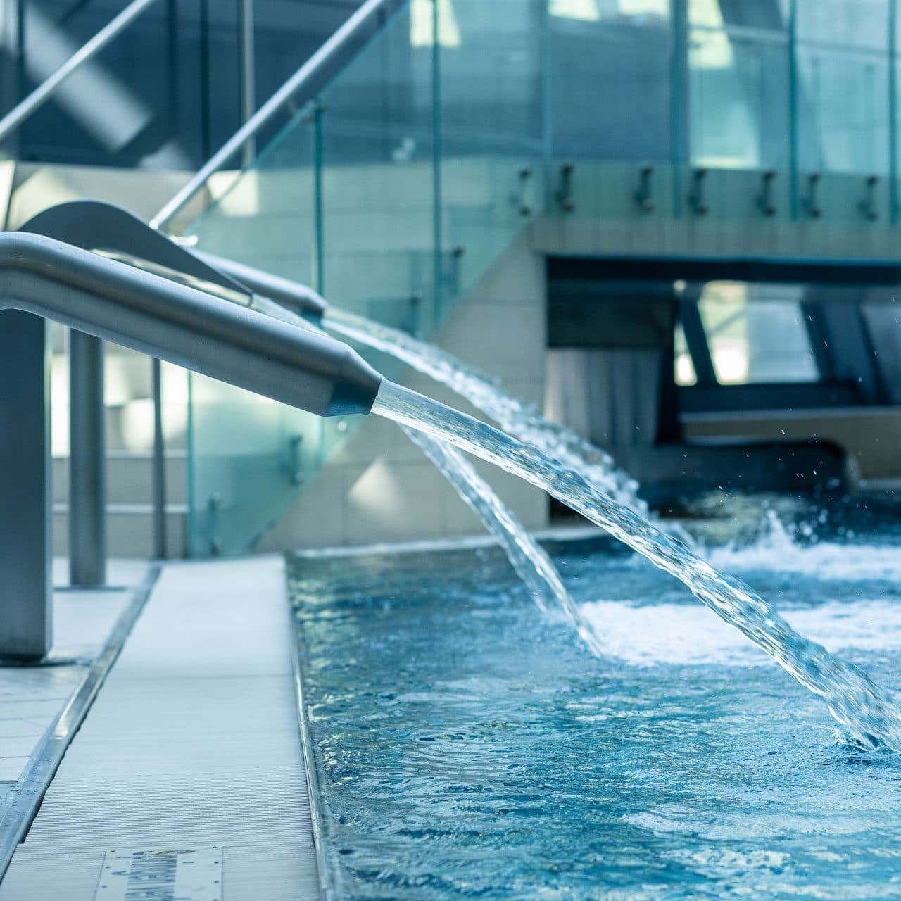 Zwei Metallspeier leiten Wasser in ein Hallenbad. Stufen führen zu einem Bereich mit Glasrand und im Hintergrund zu einer nahegelegenen Sauna.
