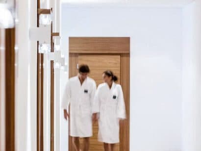 Zwei Menschen in weißen Bademänteln schlendern gemächlich einen ruhigen Flur mit Holztüren und makellos weißen Wänden entlang und rufen so die Ruhe eines luxuriösen Spa-Aufenthalts hervor.