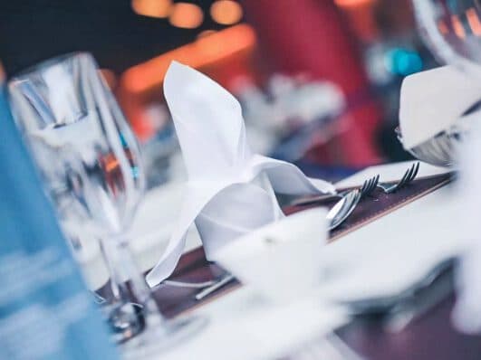 A close-up of a restaurant table setting arranged for a meeting, featuring a folded white napkin, wine glasses, forks, and a cup against a blurred background.