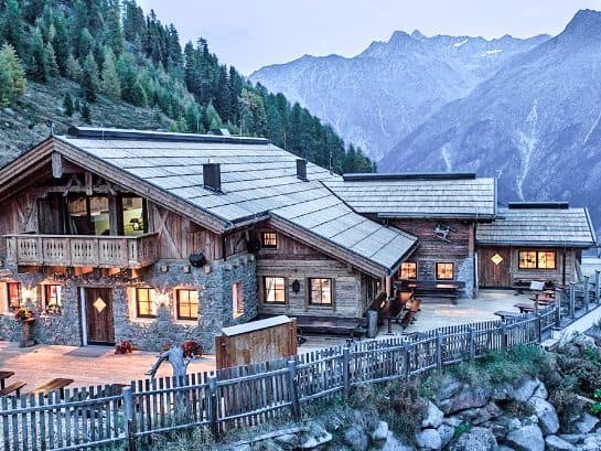A rustic mountain lodge with stone and wood architecture, designed for serene meetings, is surrounded by trees and mountains, illuminated by warm lights at dusk.