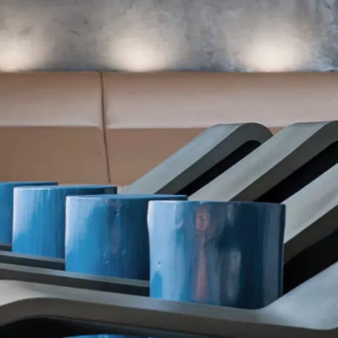 Eine Reihe moderner, blauer zylindrischer Hocker steht vor einer Wand mit stimmungsvoller Beleuchtung und ergänzt die ruhigen, beigefarbenen Sitzpolster – ein perfekter Auftakt für einen wohltuenden Saunagang.