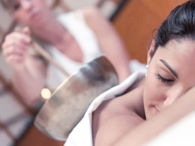 Eine Frau liegt mit geschlossenen Augen in einer ruhigen Spa-Umgebung auf der Seite, während eine andere Person sanft eine Klangschale über ihre Schultern hält.