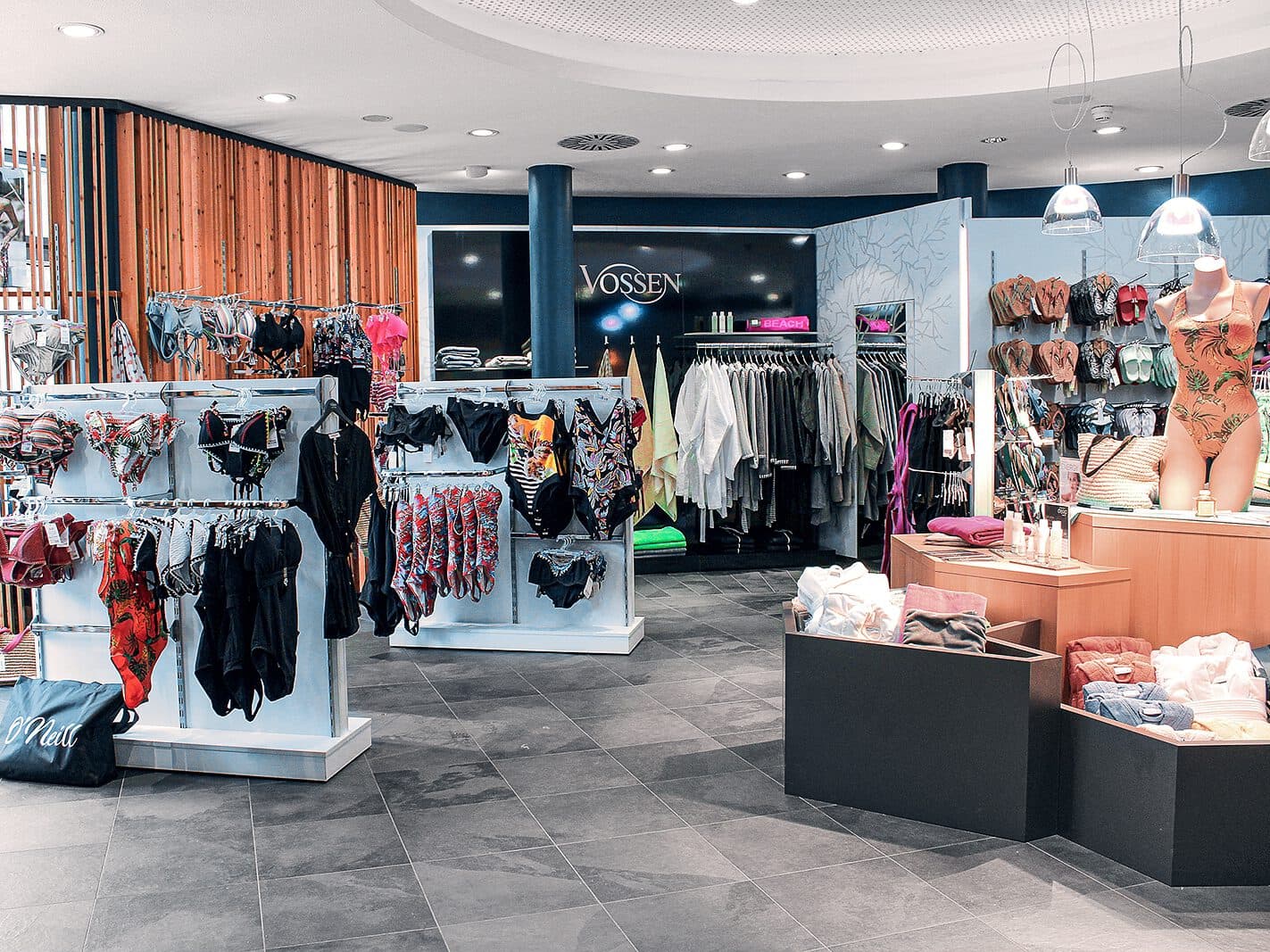 Un intérieur de magasin présentant divers maillots de bain et vêtements sur des supports et des étagères, avec des maillots de bain sur des mannequins et des vêtements pliés sur un comptoir dans un espace bien éclairé.