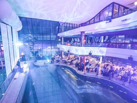The indoor pool area transforms into an elegant dining event, perfect for a meeting. Tables with guests surround the shimmering water. The room is bathed in purple lighting, accentuated by large windows and a high ceiling.