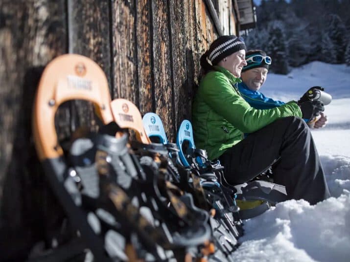Two people sit in the snow against a wooden wall, having a meeting while wearing winter gear and looking at a device. Several pairs of snowshoes are lined up beside them.