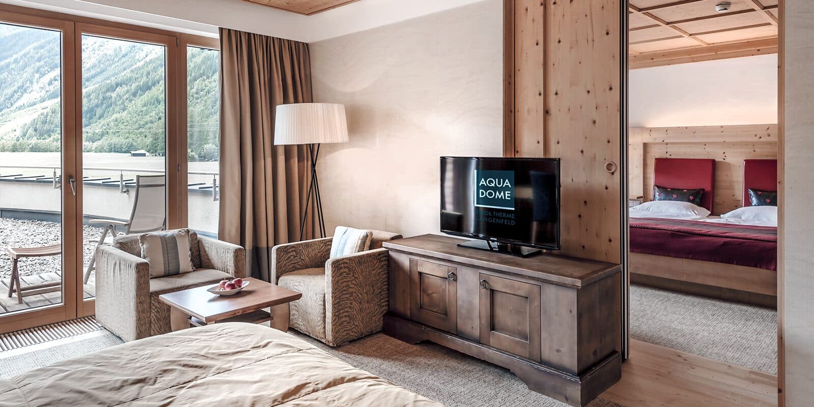 Hotelzimmer mit Doppelbett, Fernseher auf Holzständer, zwei Sesseln und einem Tisch. Großes Fenster mit Bergblick. Nebenzimmer mit zwei sichtbaren Einzelbetten. Moderne und gemütliche Inneneinrichtung.