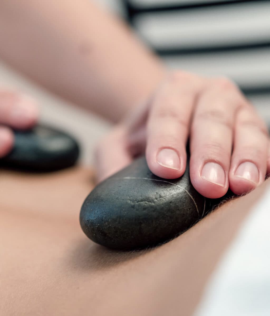 Nahaufnahme einer Person, die eine Hot-Stone-Massage erhält. Eine Hand legt glatte, schwarze Steine auf den Rücken und fördert so die Entspannung.
