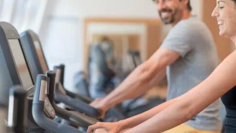 Zwei lächelnde Menschen, die in einem Fitnessstudio auf dem Heimtrainer fahren.