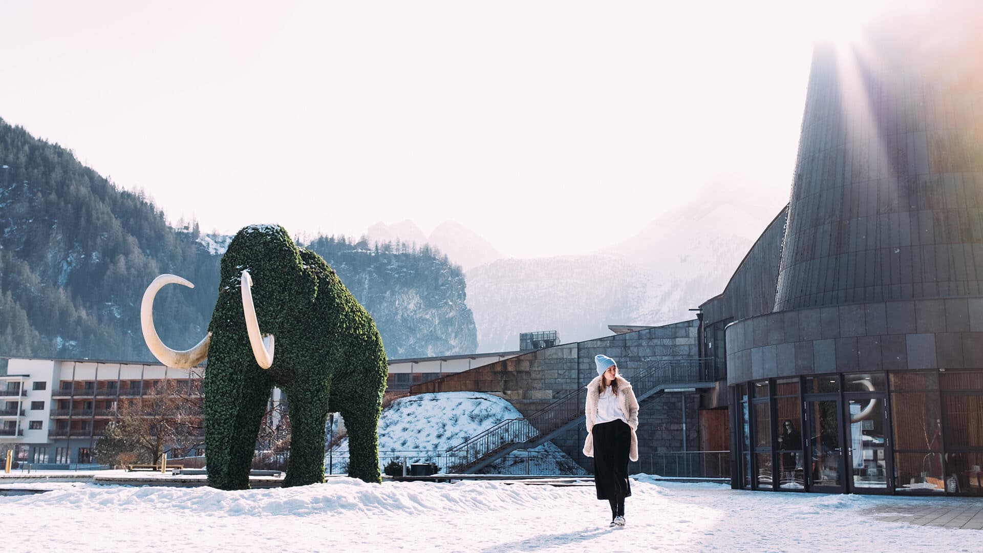Eine Person läuft auf Schnee in der Nähe von Gebäuden und einer großen grünen Mammutskulptur, im Hintergrund sind Berge unter einer hellen Sonne zu sehen.