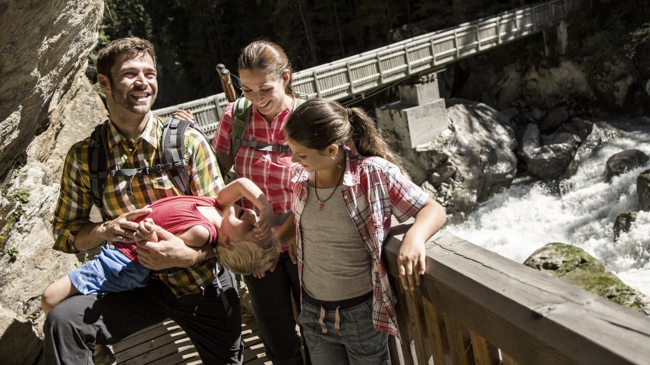 Una famiglia di quattro persone cammina vicino a un ponte su un fiume impetuoso. I genitori sono in piedi ai lati del figlio, che si sporge dalla ringhiera, mentre la figlia osserva attentamente la scena.