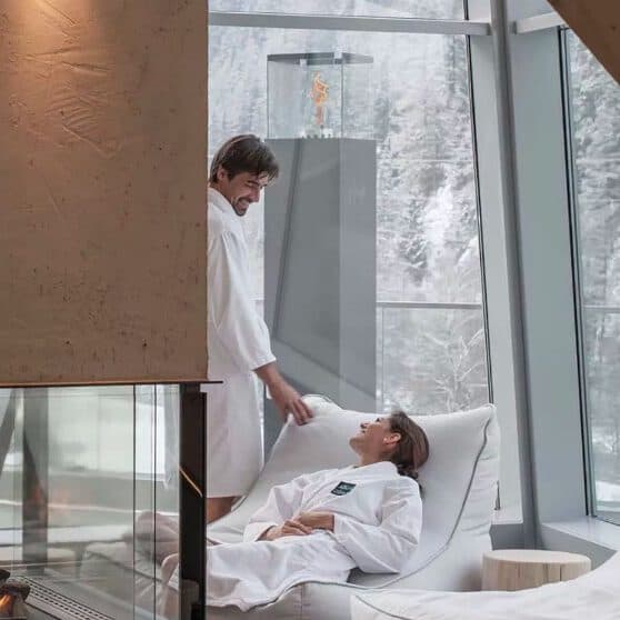 A man and woman in white robes relax by a cozy fireplace, enjoying spa-like tranquility as large windows reveal the snow-covered trees outside.