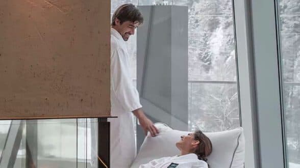 Un uomo in tunica bianca è in piedi accanto a una donna sdraiata, anch'essa in tunica bianca, in un interno da cui si può ammirare un paesaggio innevato attraverso grandi finestre.