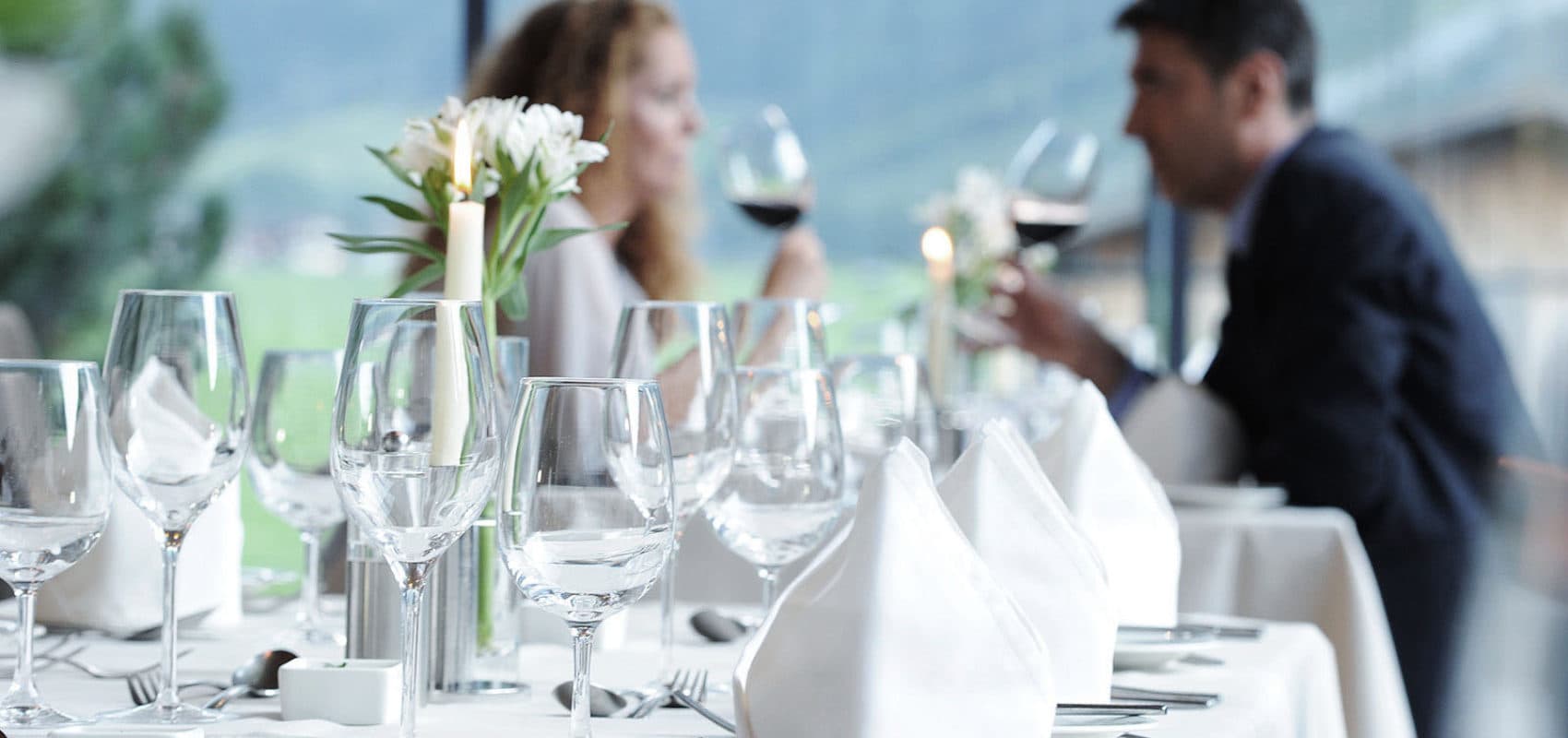 Ein mit leeren Gläsern und gefalteten Servietten gedeckter Tisch, im Hintergrund genießt ein Paar in einem Restaurant Wein.