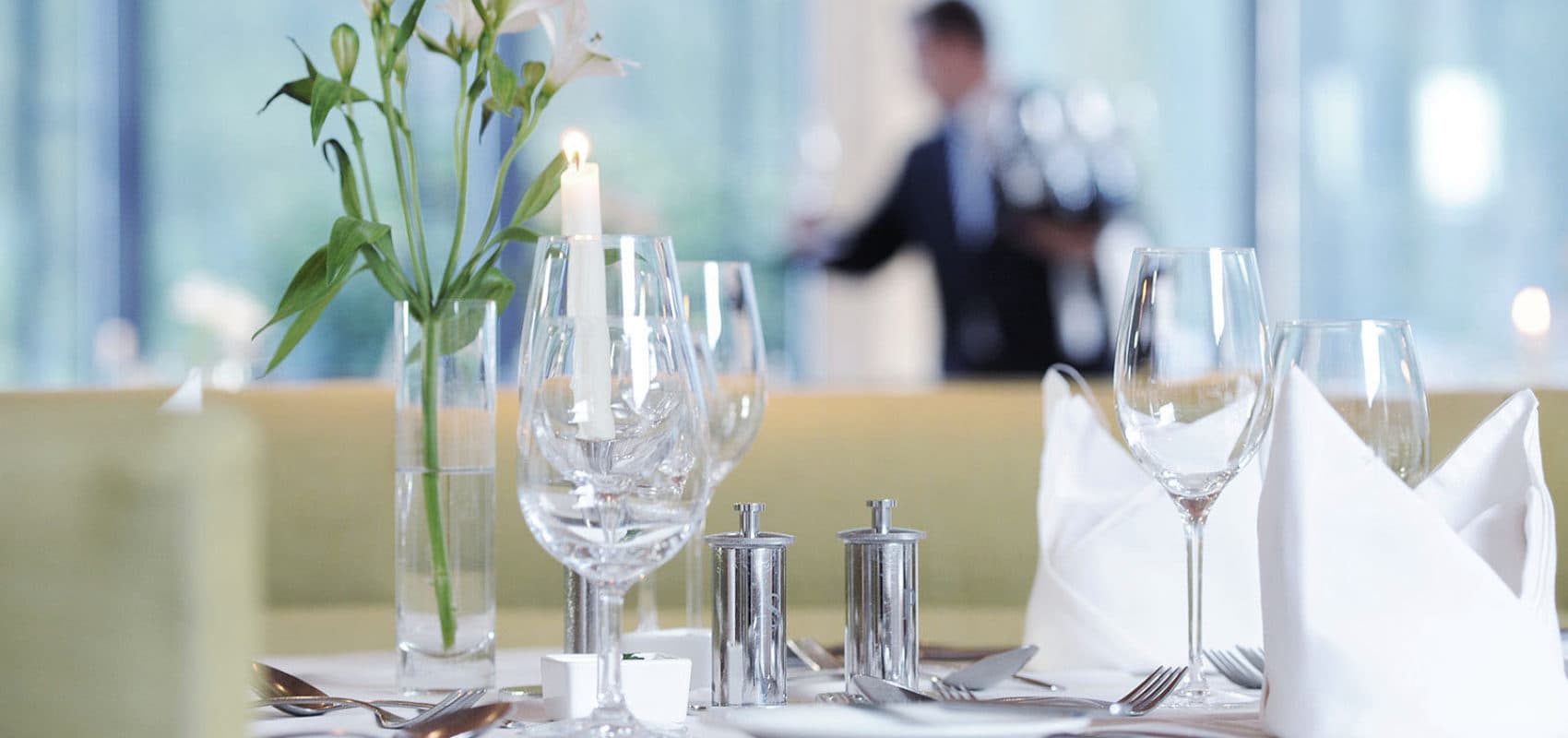 Ein Esstisch mit Gläsern, Besteck, Salz- und Pfefferstreuern, weißen Servietten und einer Blumenvase. Im Hintergrund trägt eine Person ein Tablett mit Gläsern.