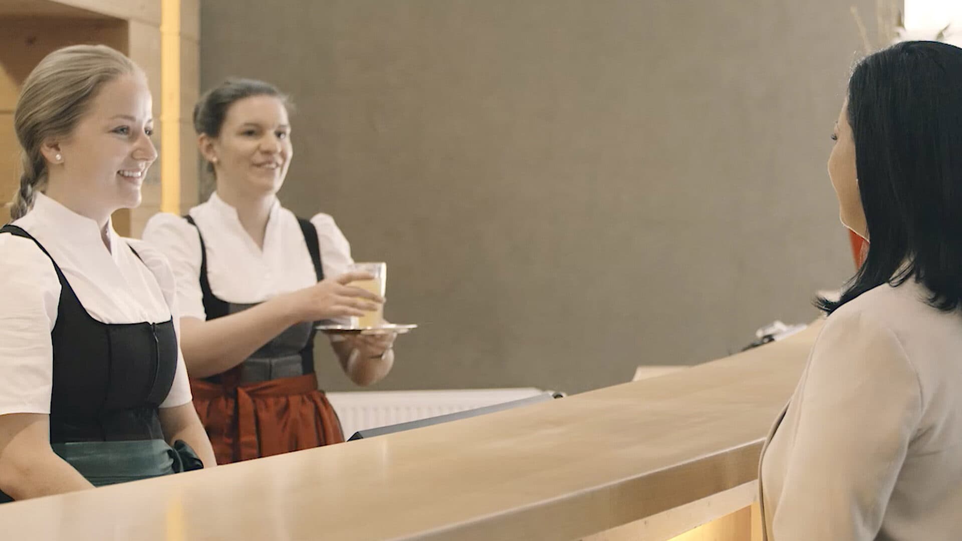 Zwei Frauen in traditioneller Kleidung servieren einem Kunden Getränke über eine Holztheke in einem in neutralen Farbtönen gehaltenen Raum.