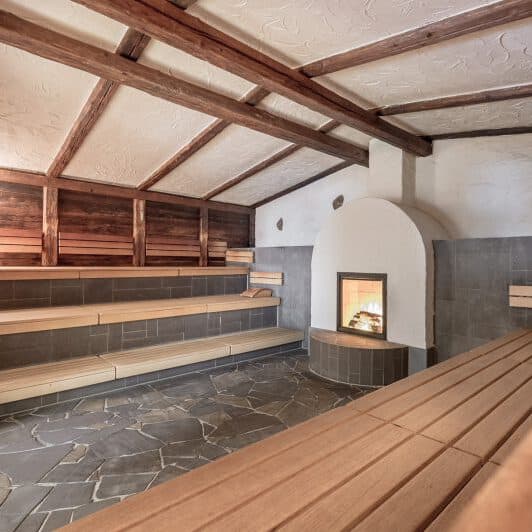 Сауна с деревянными панелями, полом из каменной плитки, деревянными скамейками и зажженным камином, установленным в белой нише с одной стороны.