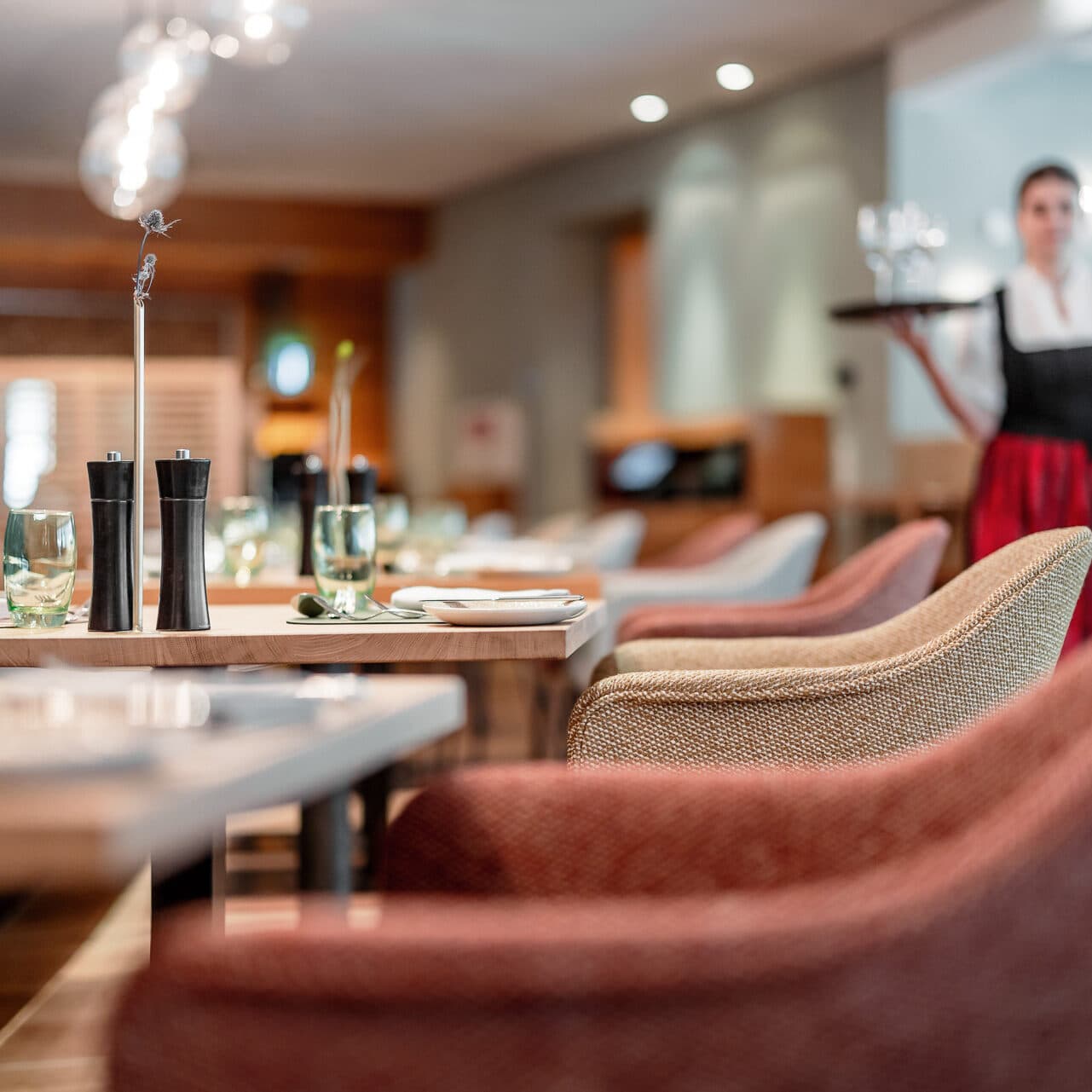 Ein Restaurant im Galerie-Stil mit gepolsterten Stühlen umgibt einen mit Weingläsern und einer Kerze geschmückten Tisch und strahlt Eleganz aus. Im Hintergrund trägt ein Kellner anmutig ein Tablett und unterstreicht das künstlerische Ambiente.