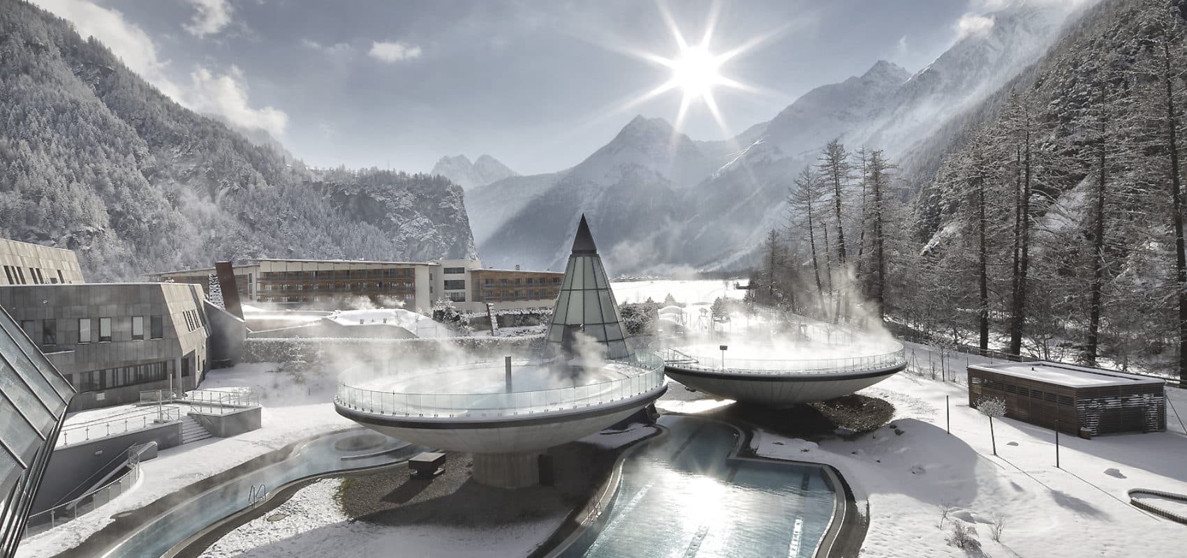 Erleben Sie einen Winterurlaub in unserem Bergresort, wo dampfende Außenpools zwischen schneebedeckten Bäumen und Gebäuden liegen und die Sonne strahlt.