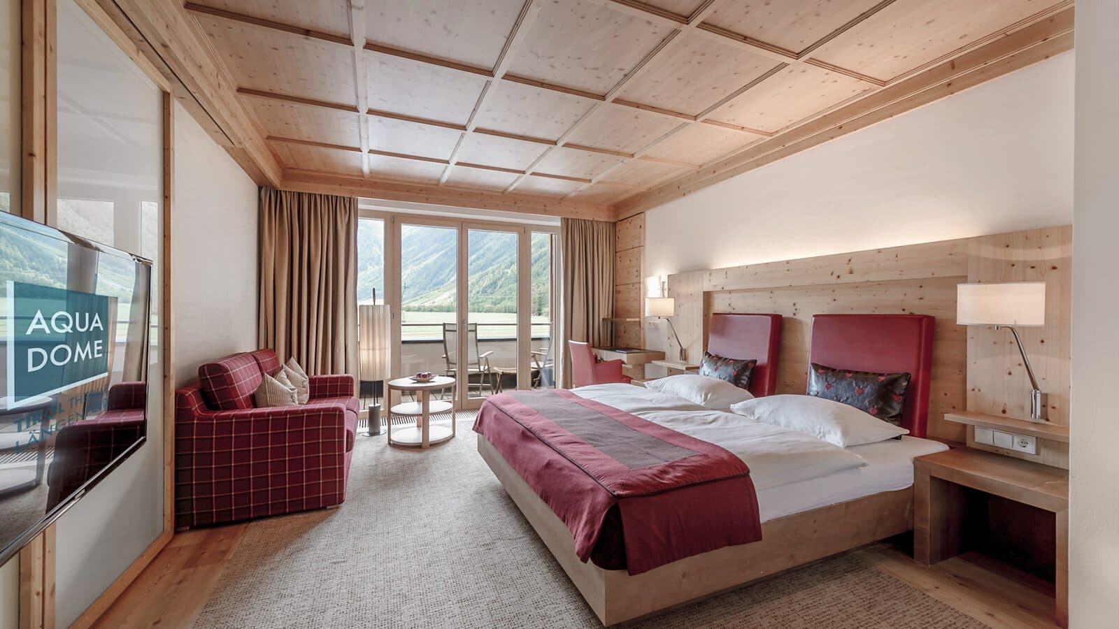 Das Hotelzimmer besticht durch ein großes Bett und rote Akzente, ein bequemes Sofa und einen Fernseher mit dem Titel „Aqua Dome“. Durch die Glastüren gelangen Sie auf den Balkon und genießen den atemberaubenden Blick auf die Berge – ein wahres Kunstwerk der Natur.