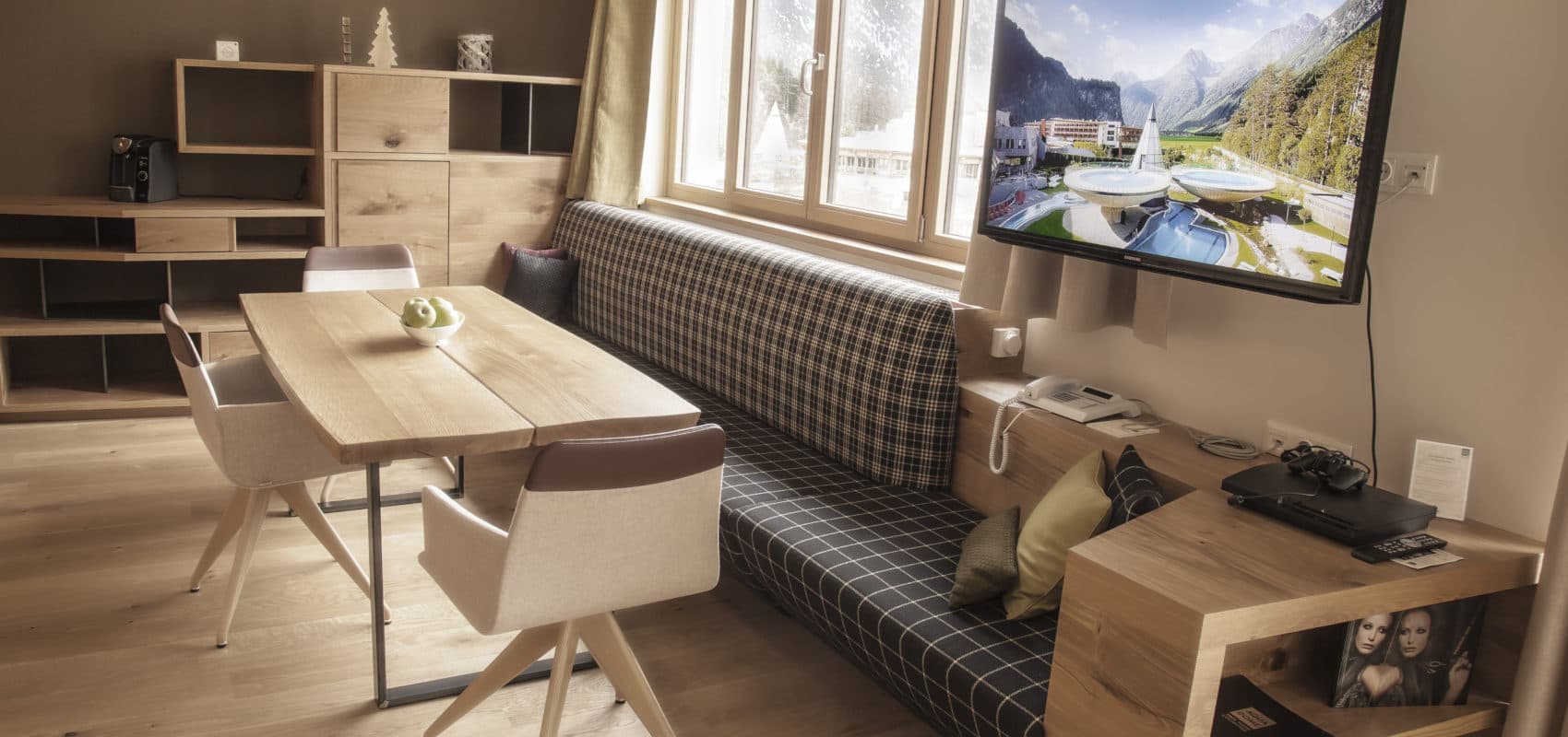 Modernes Wohnzimmer mit Holzboden, Tisch und Stühlen, kariertem Sofa und einem an der Wand montierten Fernseher mit malerischer Aussicht, die an einen Winterurlaub erinnert. Im Hintergrund sind Regal und Dekoration zu sehen.