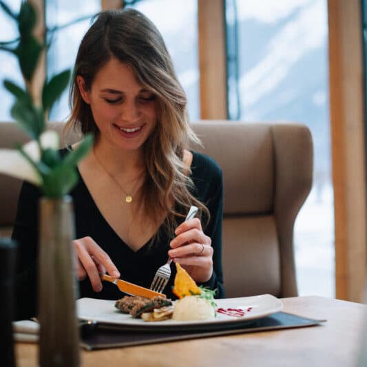 Женщина в черном топе сидит за столиком в ресторане, улыбаясь и нарезая еду на тарелке. На заднем плане — окно с заснеженным видом.
