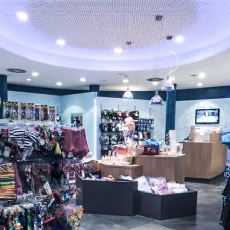 Eine hell erleuchtete Boutique ähnelt einer fesselnden Galerie, in der auf Ständern und Tischen verschiedene farbenfrohe Kleidungsstücke und Accessoires präsentiert werden, während im Hintergrund in der Nähe der Kasse weitere einzigartige Artikel zu finden sind.