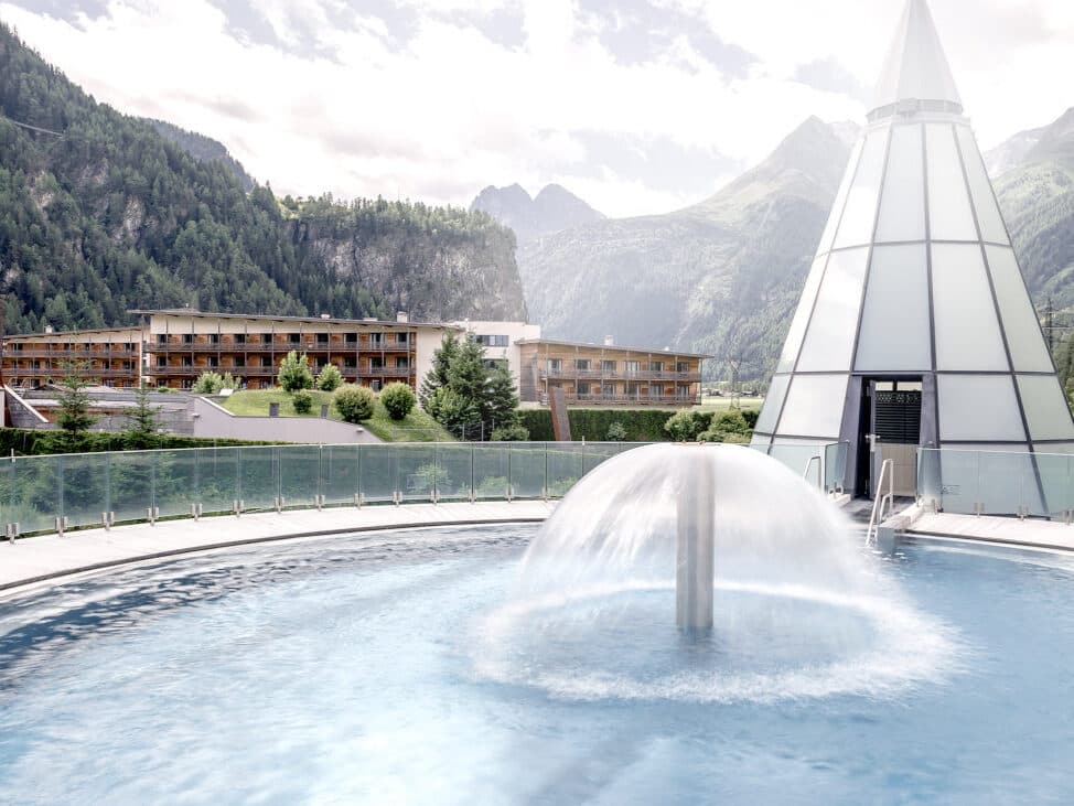 Eine moderne Glaspyramide und ein großer Brunnen in einem runden Becken bilden das perfekte Herzstück für einen erholsamen Thermenurlaub, alles vor der Kulisse majestätischer Berge und eines mehrstöckigen Gebäudes.