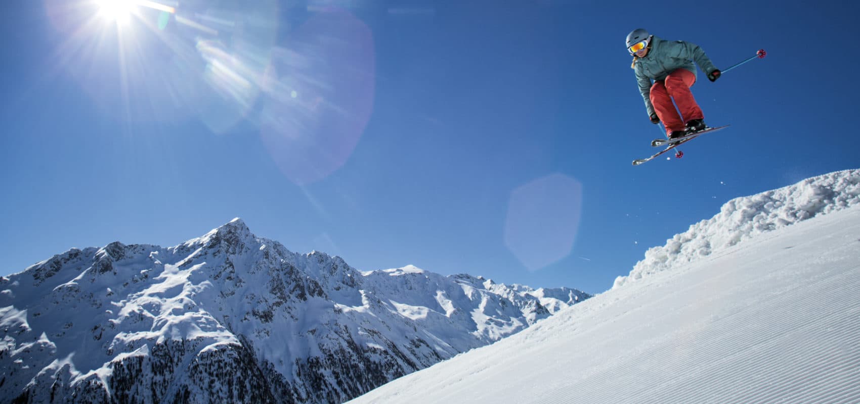 Ein Skifahrer macht einen Sprung in der Luft auf einem sonnigen Winterurlaubshang mit schneebedeckten Bergen im Hintergrund.